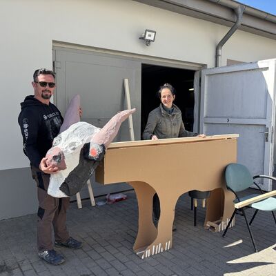 Ein Mann mit schwarzem Hoodie, dunklen Haaren udn Sonnenbrille tr&auml;gt einen gebastelten und bemalten Kuhkopf auf Pappmach&eacute;. Im Hintergrund eine Frau, die vor einer ge&ouml;ffneten Garagent&uuml;r steht und einen gebastelten, unbemalten Kuhk&ouml;rper aus Pappe h&auml;lt.